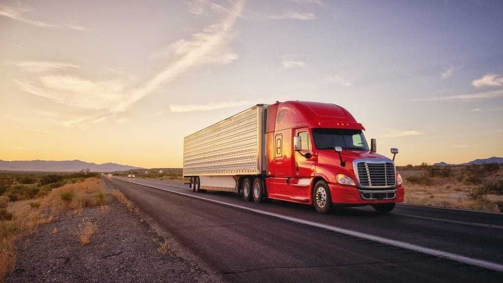 Red semi-truck with trailer driving on a highway through a desert landscape at dusk.