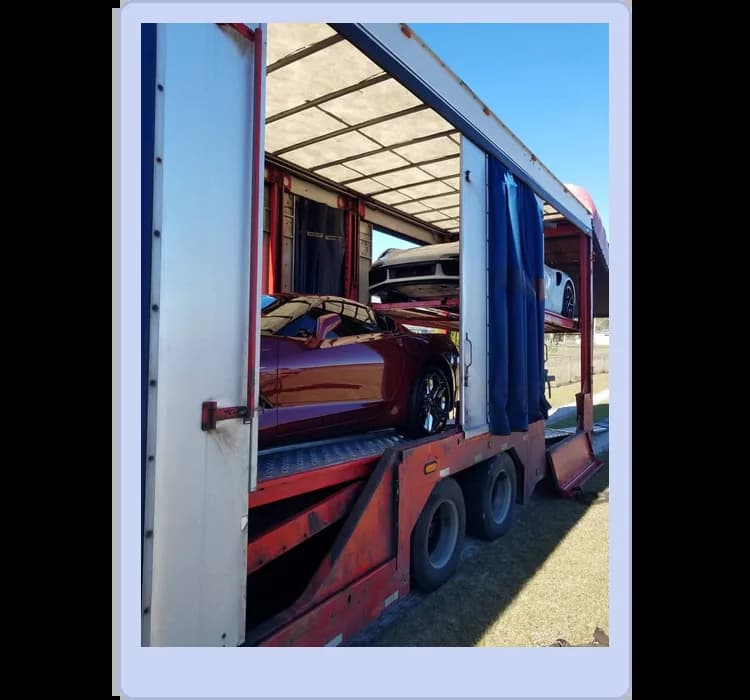 Two sports cars are being transported inside an open-sided red car carrier trailer on a sunny day.
