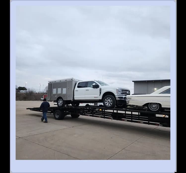 A man walks near a car hauler trailer loaded with a white utility truck and a white classic car in an industrial lot.