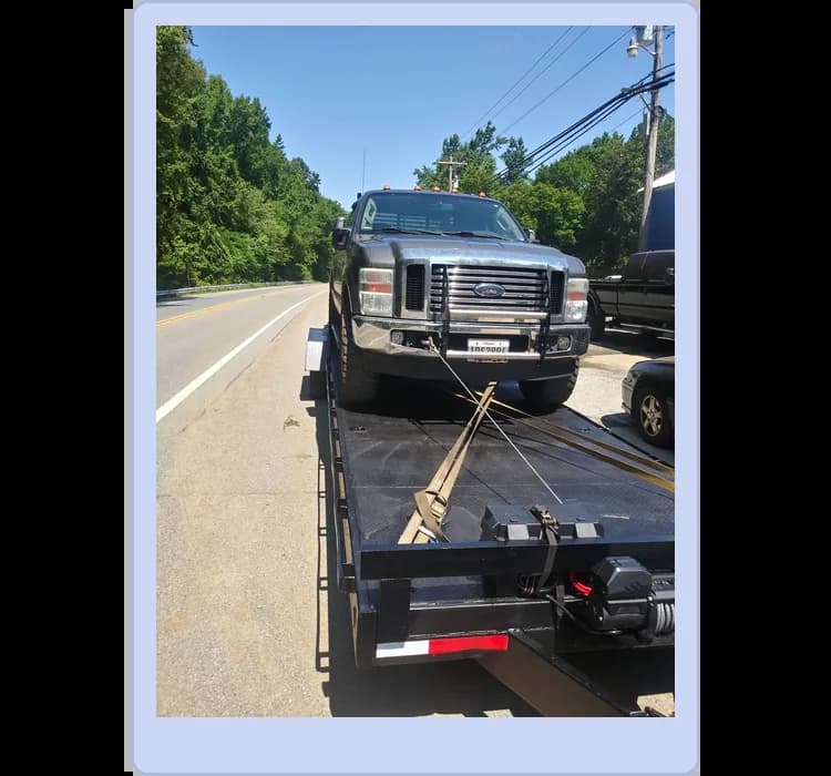 A black pickup truck is secured on a flatbed tow trailer by straps, parked on the side of a two-lane road on a sunny day.