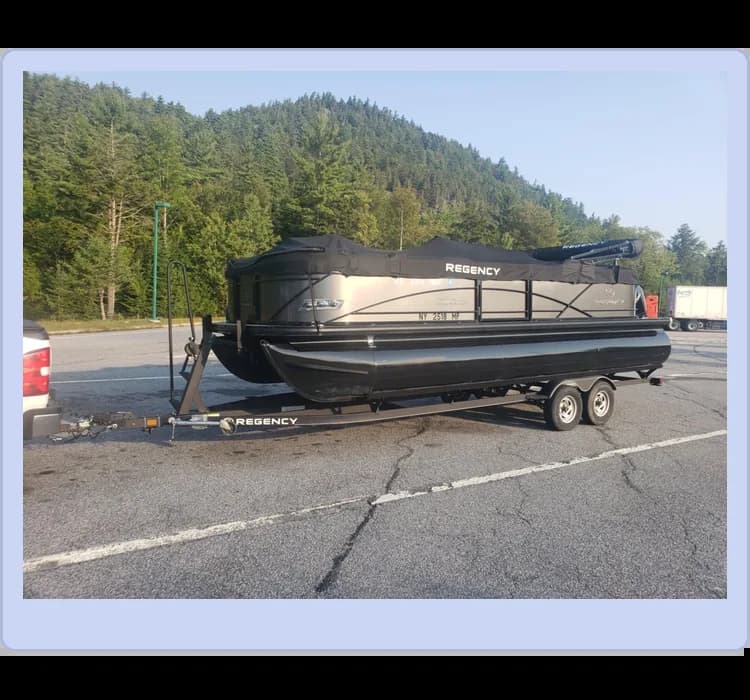 A black pontoon boat on a trailer parked in a lot, surrounded by trees and hills in the background.