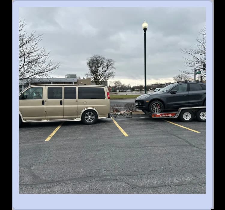 A beige van is parked in a lot, towing a black SUV on a U-Haul trailer. The sky is overcast and trees are bare in the background.