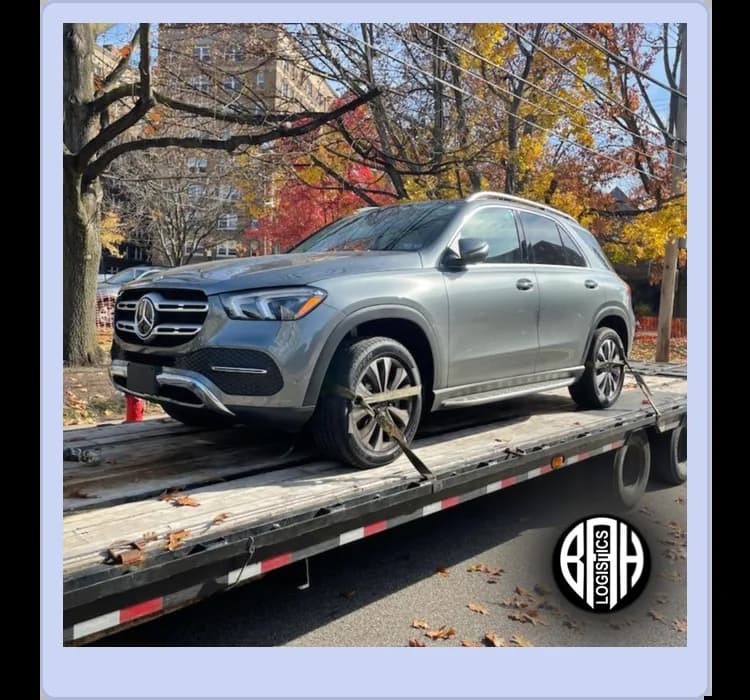 A silver Mercedes-Benz SUV is secured on a flatbed trailer for transport, with trees and buildings visible in the background.
