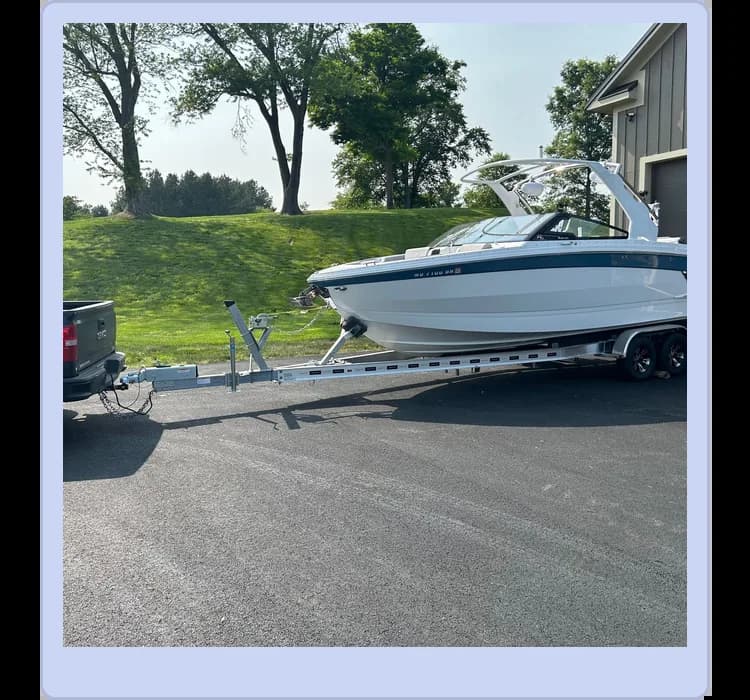 A boat on a trailer is attached to a pickup truck on a paved driveway near a house, with grass and trees in the background.