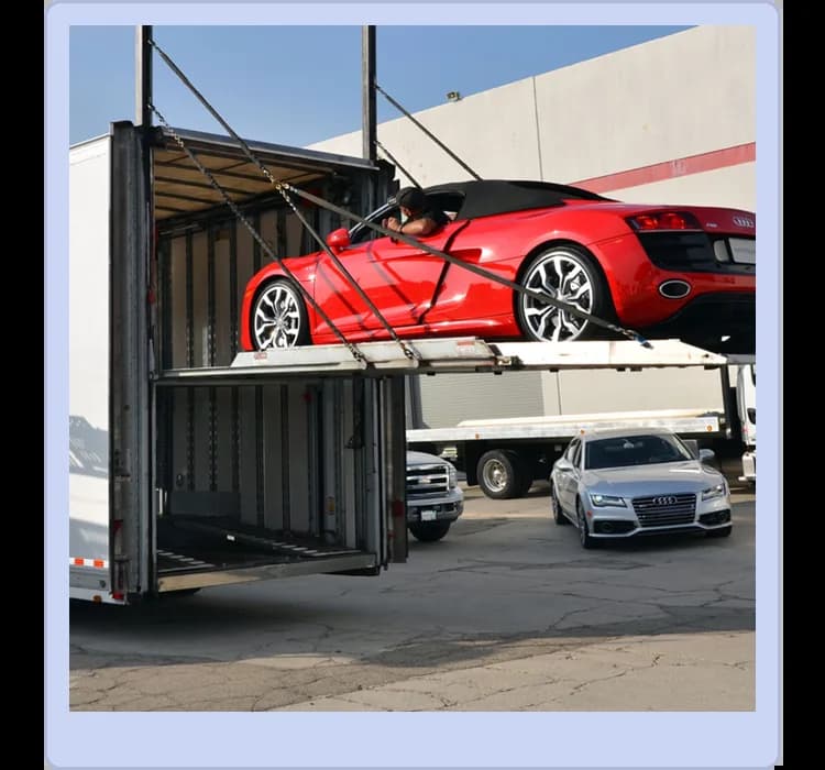 A red convertible sports car is being loaded into a large enclosed car carrier truck, with another car and trucks visible in the background.