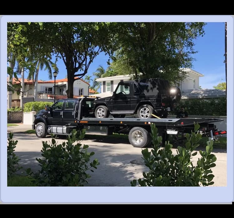 A black Mercedes-Benz G-Class SUV is being transported on a flatbed tow truck in a residential neighborhood with trees and houses in the background.