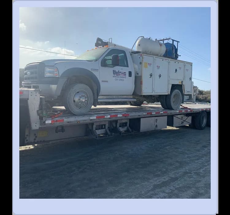 A white utility truck with equipment mounted on its back is loaded onto a flatbed trailer for transport on a dirt road.