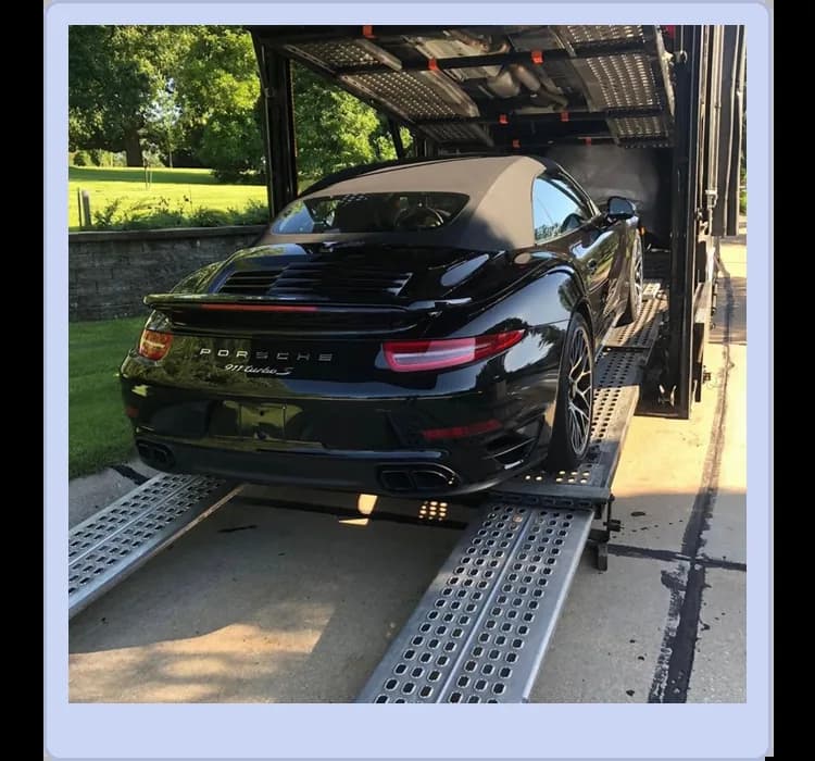 A black Porsche 911 Turbo convertible is being loaded onto a car transport trailer outdoors on a sunny day.