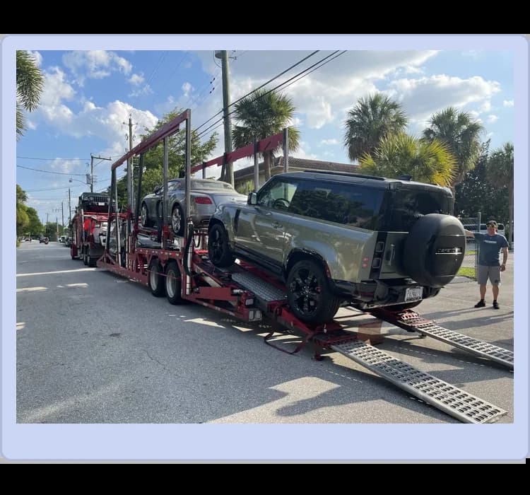 A car carrier trailer loaded with several vehicles, including a green Land Rover Defender, parked on a sunny street.