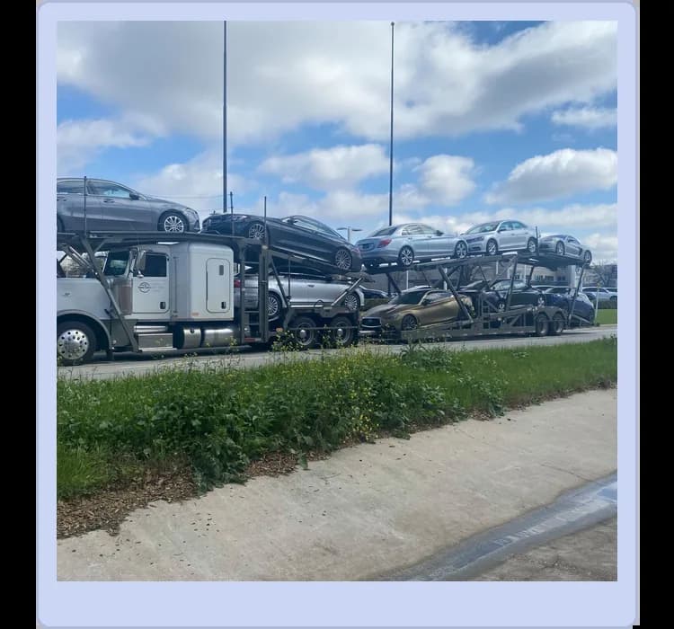 A car carrier truck is transporting several cars on a highway, with blue sky and clouds in the background.