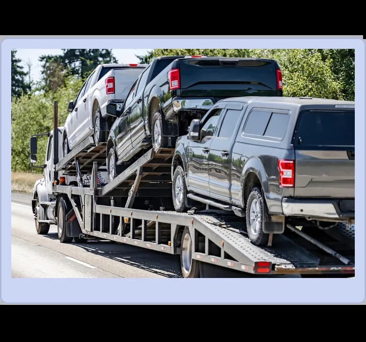 A car carrier truck transports three pickup trucks on an open trailer along a road, with trees and greenery in the background.