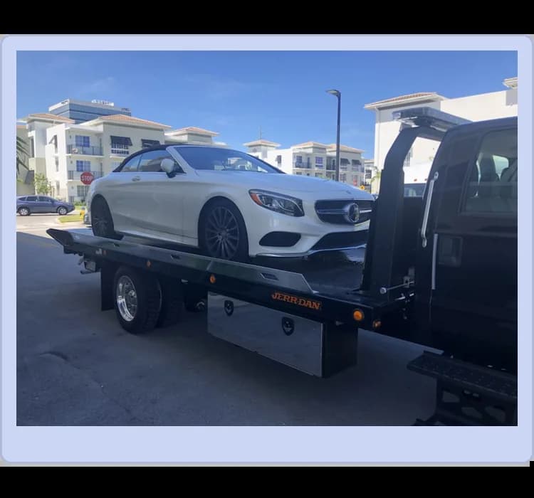 A white Mercedes-Benz convertible is being transported on the flatbed of a black tow truck in front of modern buildings under a clear blue sky.