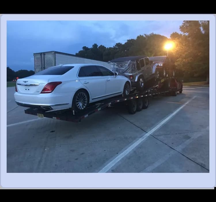 A car transporter trailer parked in a lot carries a white sedan and a gray SUV wrapped in protective plastic.