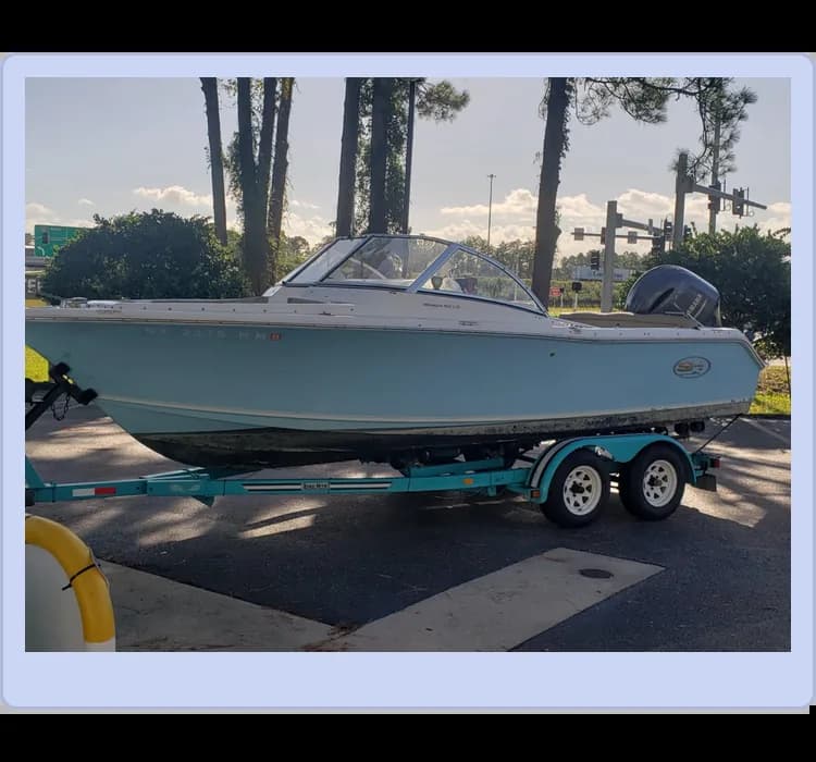 A light blue motorboat on a trailer with dual axles is parked on a paved area. Trees and a road sign are visible in the background.