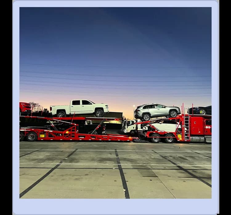 A red car carrier trailer loaded with several vehicles, including a white pickup truck and a silver SUV, parked on a lot at dusk.