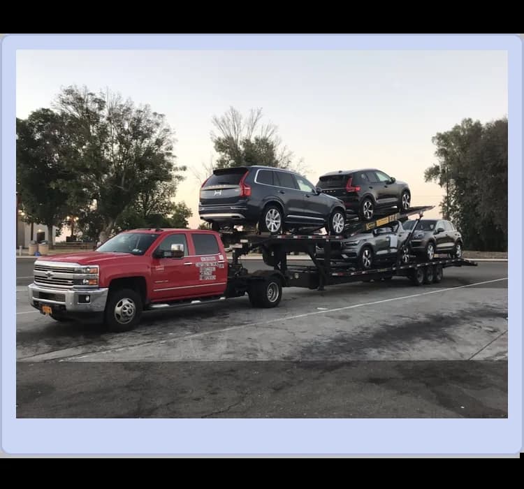 A red pickup truck towing a multi-level car trailer loaded with six vehicles, parked in an outdoor lot with trees in the background.