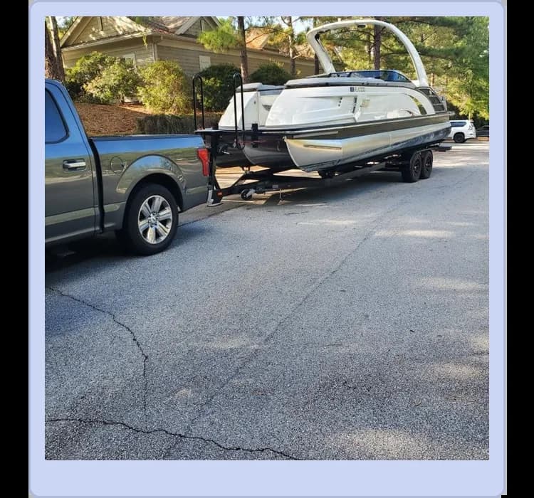A truck is parked on a residential street towing a large boat on a trailer. Houses and trees are visible in the background.
