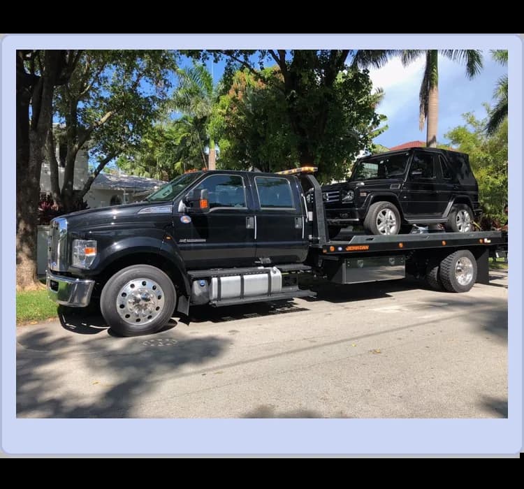 A black tow truck is parked on a street, carrying a black SUV on its flatbed. Trees and houses are visible in the background.
