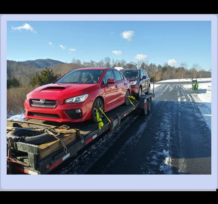 A red car and a gray SUV are strapped onto a flatbed trailer parked on a snowy road with trees and hills in the background.