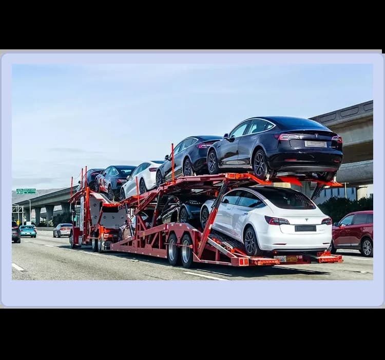 A red car carrier trailer transports several Tesla vehicles on a multi-lane highway under a clear sky.