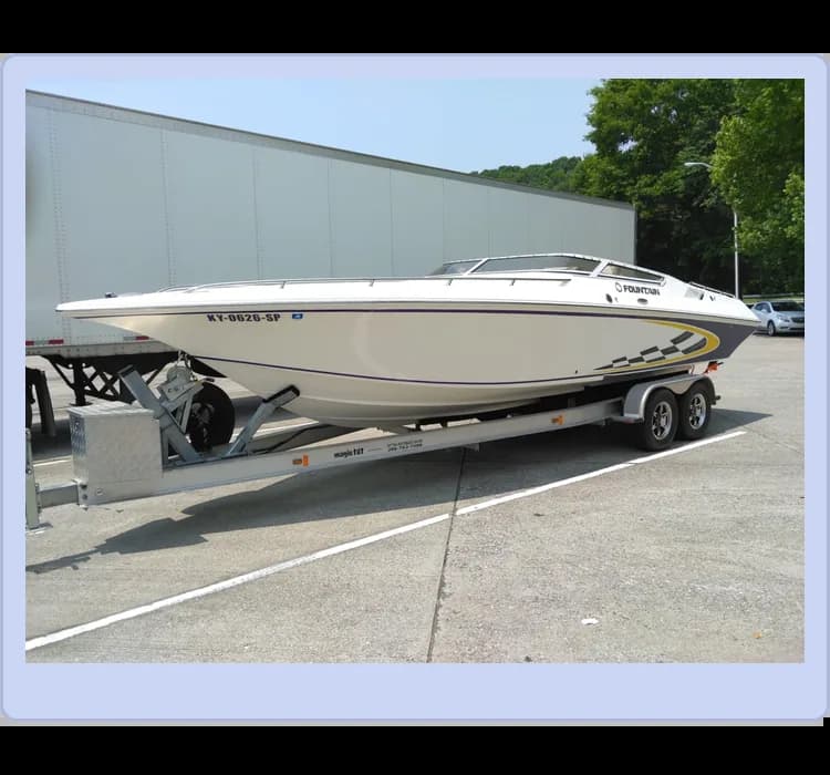 A white powerboat with racing stripes sits on a dual-axle trailer in a paved parking lot, near a large white truck. Trees are visible in the background.