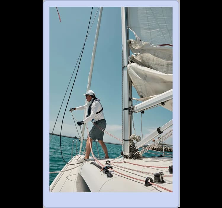 A person stands on a sailboat, adjusting the ropes with one hand while holding the side of the boat with the other. The sky is clear and the water is calm.
