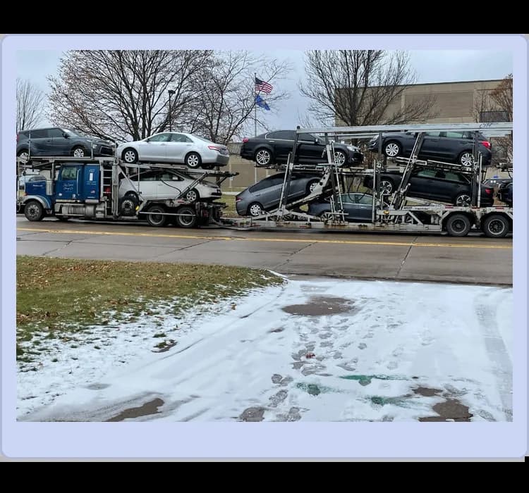 A car carrier truck transports multiple sedans on a snowy roadside, with bare trees and a building in the background.