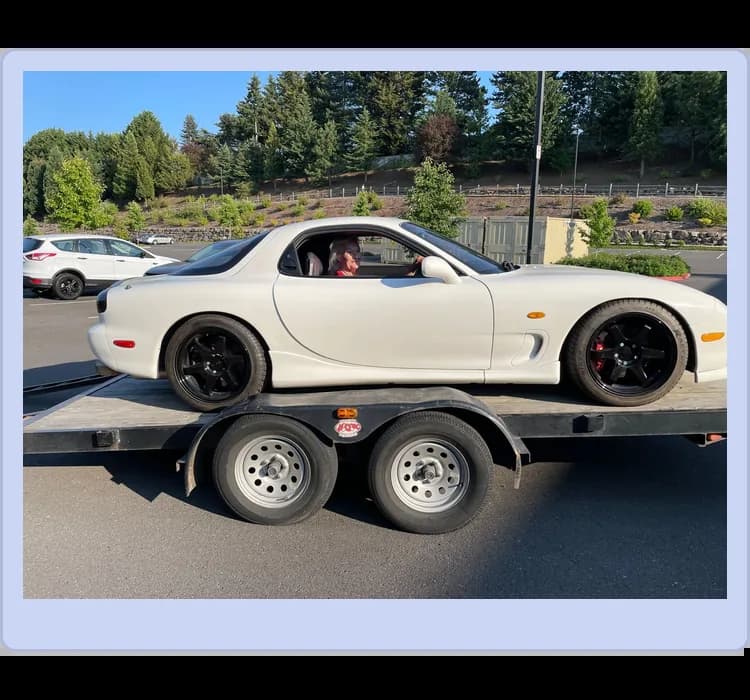 A white sports car with black wheels is parked on a trailer, with a person sitting inside. Trees and a parking lot are visible in the background.