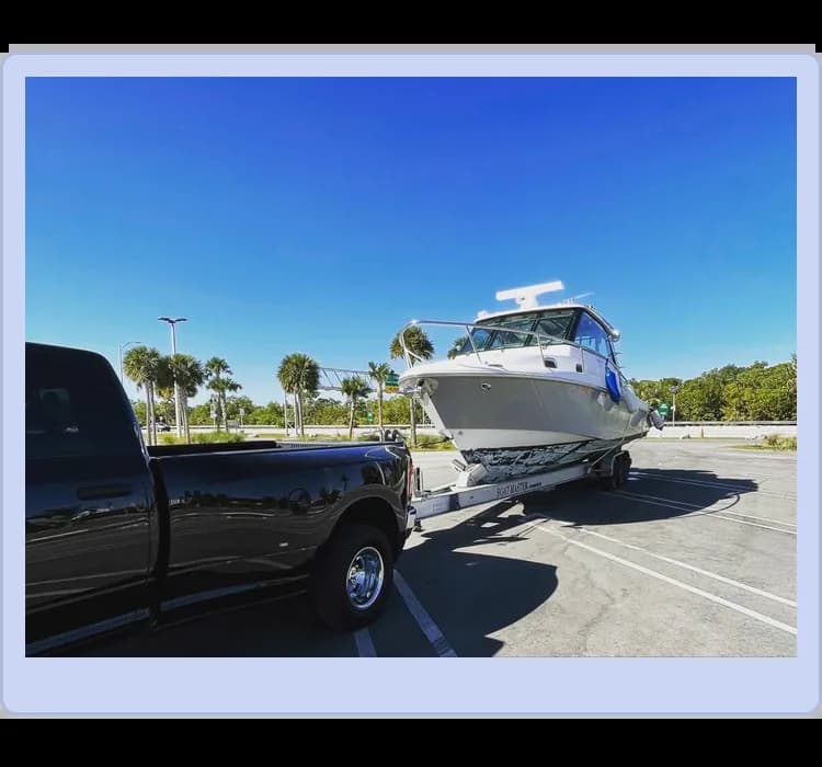 A black pickup truck is towing a boat on a trailer through an empty parking lot on a clear, sunny day. Palm trees and a bright blue sky are in the background.