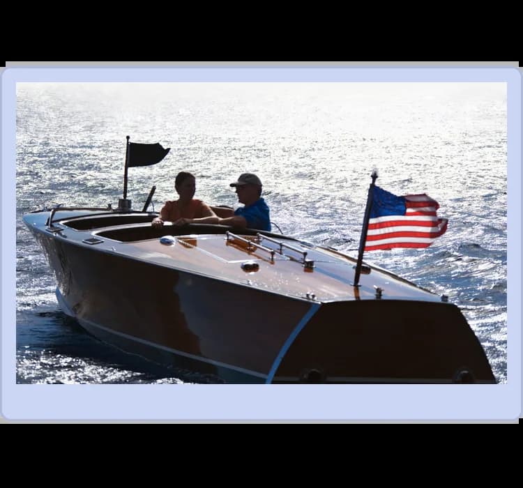 Two people in a wooden speedboat on the water, with the American flag on display at the rear.