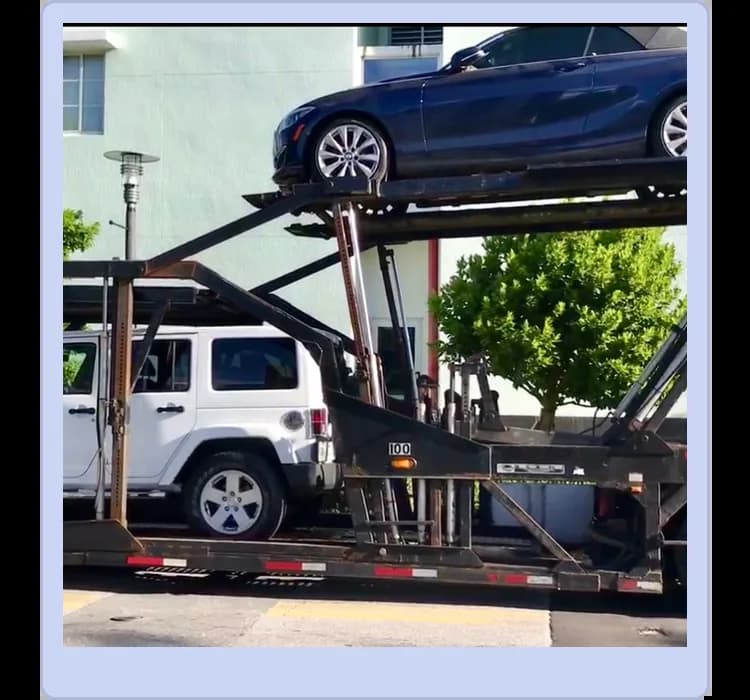 A car carrier trailer transports a blue sedan on the upper level and a white SUV on the lower level, with trees and a building in the background.