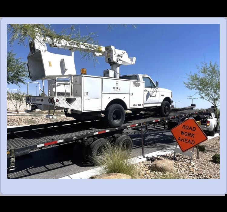 A utility bucket truck is loaded onto a flatbed trailer next to a "Road Work Ahead" sign on a sunny day.