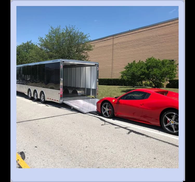 A red sports car is parked near an open car trailer on a paved road, with the trailer ramp lowered and trees and a large building in the background.
