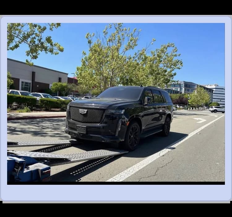 A black SUV is being loaded onto a tow truck on a sunny day, with buildings, parked cars, and trees visible in the background.
