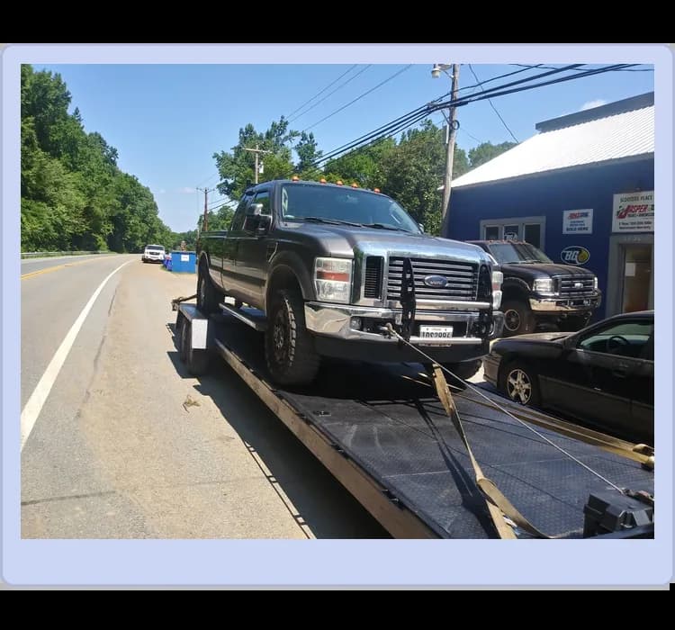 A black Ford pickup truck is secured on a flatbed trailer beside a road, with another car and a blue building in the background.