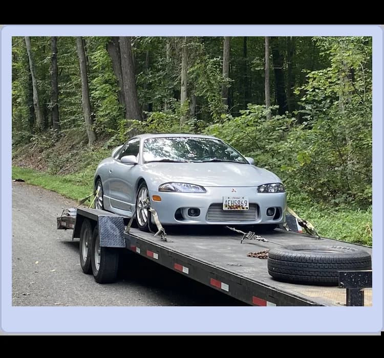 A silver Mitsubishi Eclipse is secured on a flatbed trailer parked on a wooded roadside, with a spare tire and tools nearby.
