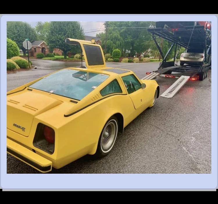 A yellow sports car with a gullwing door open is parked near a car carrier ramp on a wet street, with another car visible on the transporter.