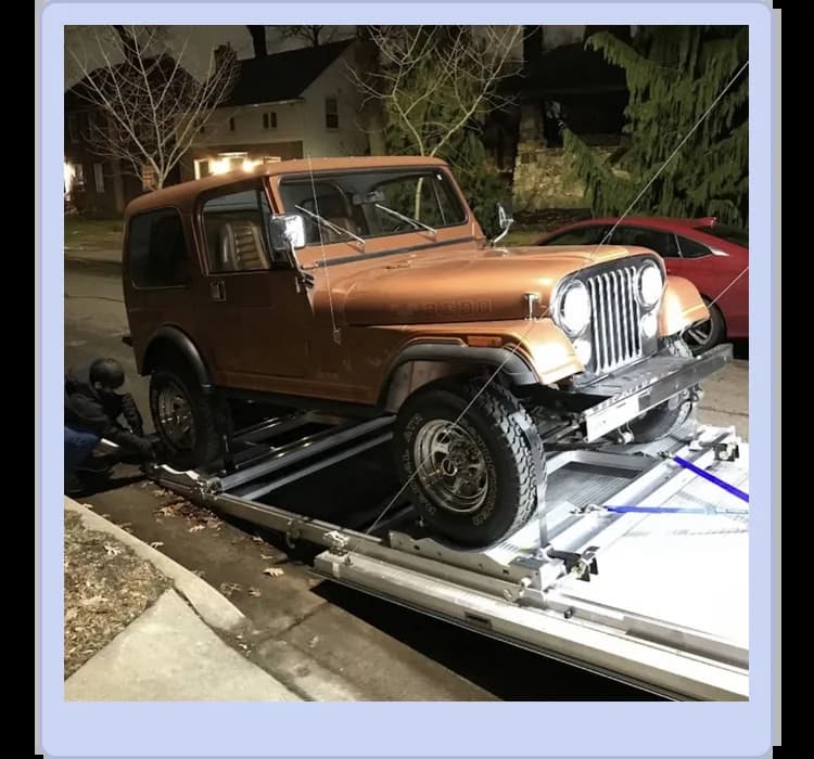 A brown vintage Jeep is being loaded onto a flatbed tow truck at night on a residential street.