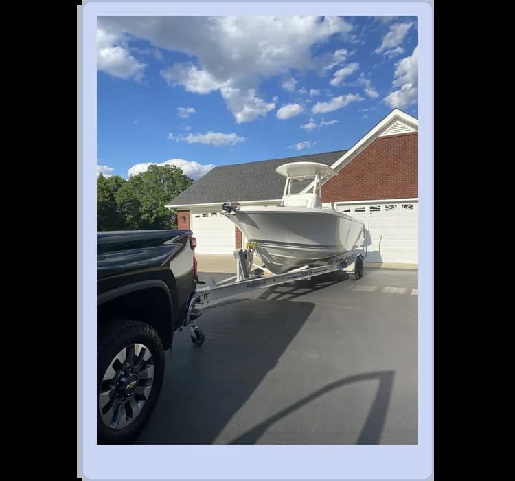 A white boat on a trailer is hitched to a black pickup truck, parked in front of a house with three garage doors. The sky is partly cloudy.