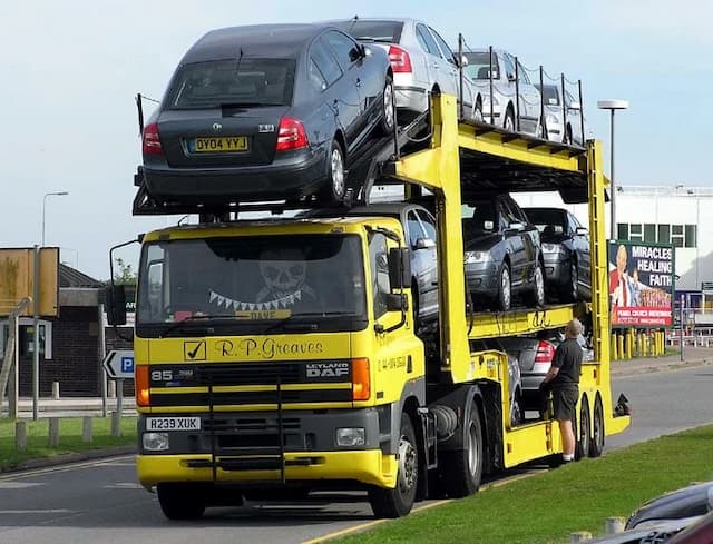 A yellow car transporter truck loaded with eight cars parked on a roadside with buildings in the background.
