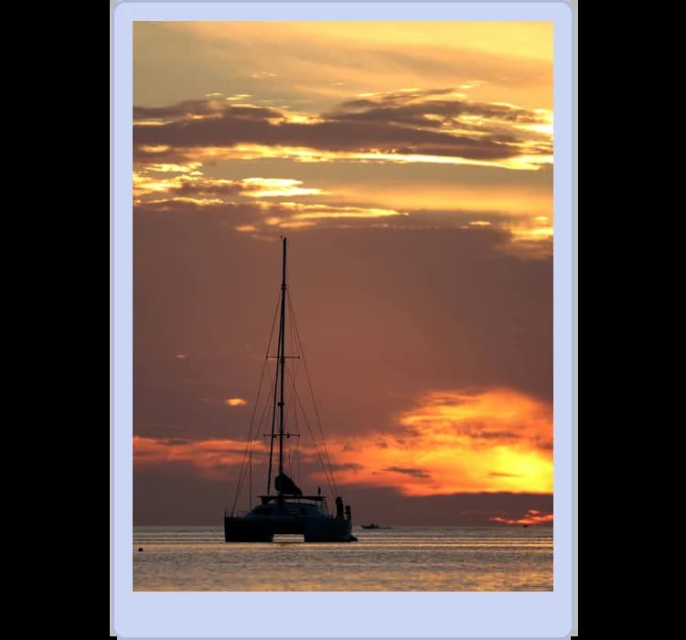 A sailboat is silhouetted against an orange and yellow sunset over calm waters, with clouds scattered across the sky.
