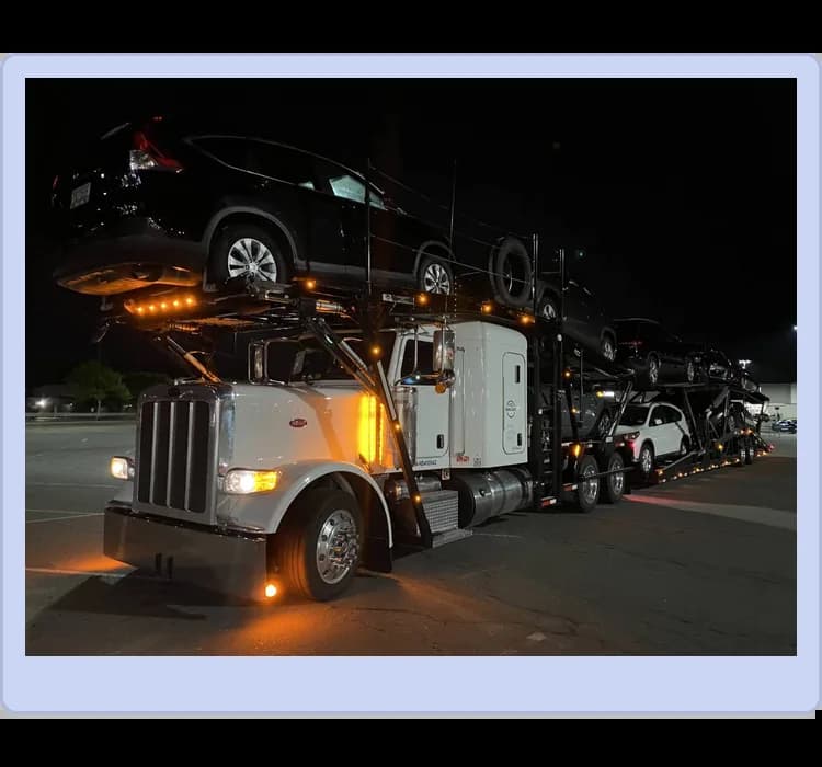 A car carrier truck loaded with multiple vehicles is parked in a lot at night, illuminated by its lights.