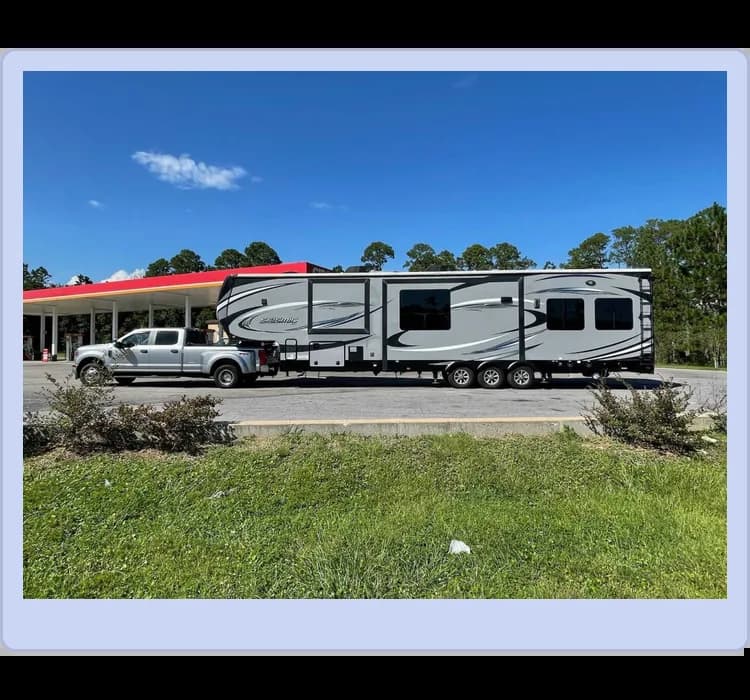 A silver pickup truck is towing a large, multi-axle fifth-wheel RV trailer near a gas station with a red roof and trees in the background.