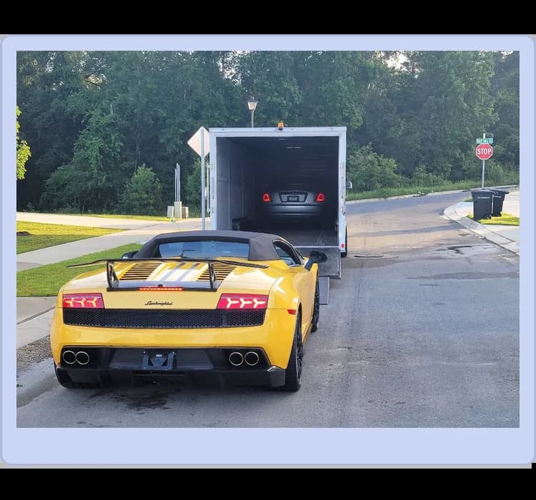 A yellow Lamborghini convertible is parked on the street behind an open car trailer, with another car visible inside the trailer. Trees and a stop sign are in the background.