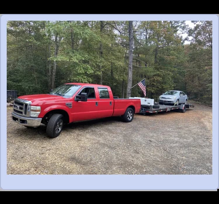 A red pickup truck towing a flatbed trailer carrying a silver car and a flag, parked on a gravel surface near trees.