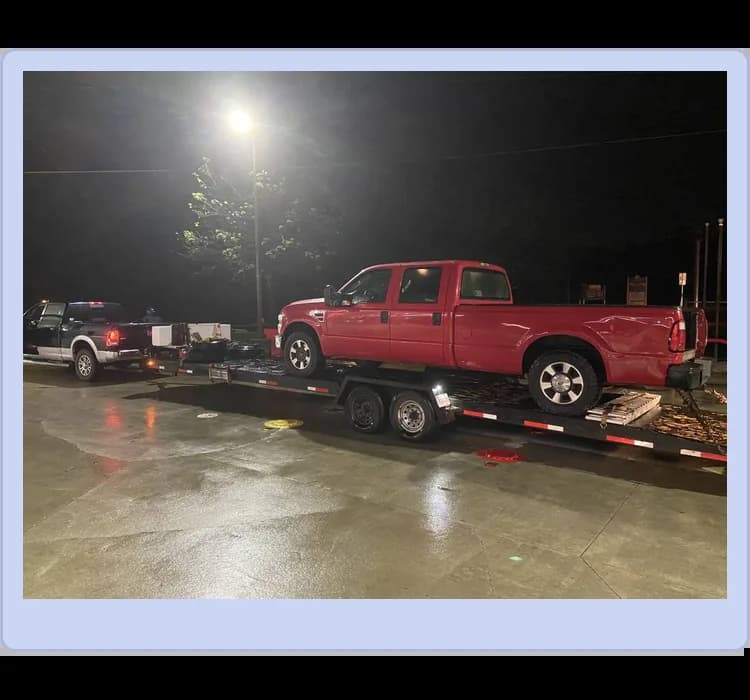 A red pickup truck is loaded onto a flatbed trailer, which is hitched to a black pickup truck at night in a well-lit area.