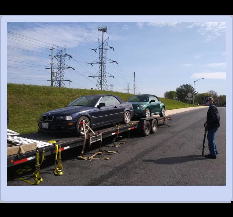 A flatbed trailer carries a black BMW convertible and a green Volkswagen Beetle; a person stands nearby on the road.