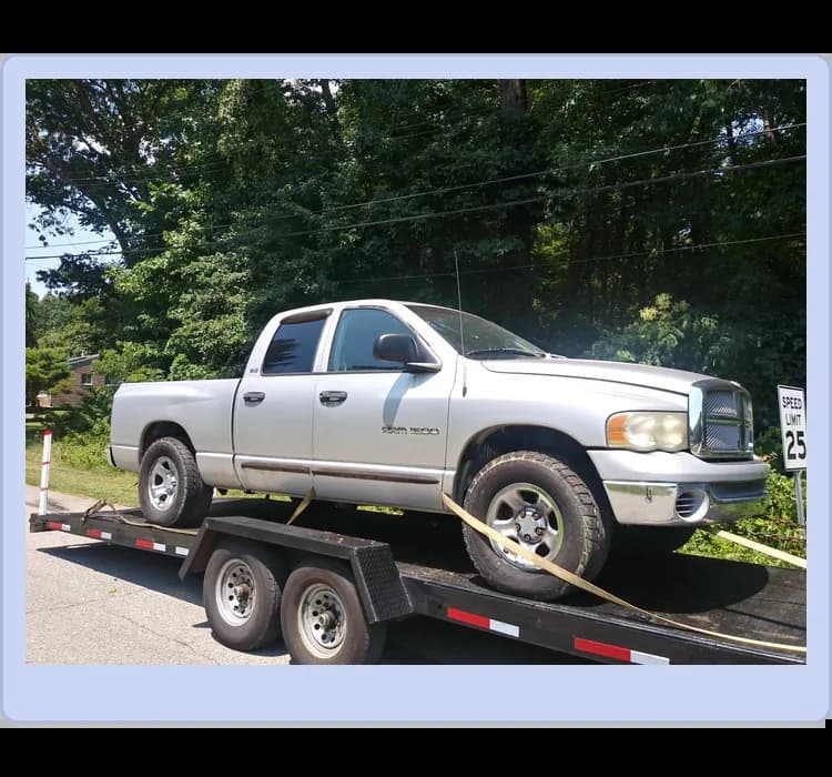 A silver Dodge Ram 1500 pickup truck is secured on a black flatbed trailer parked near a 25 mph speed limit sign and trees.