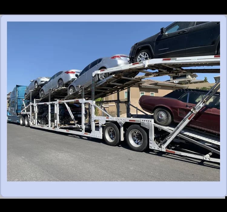 A car carrier trailer transports multiple vehicles, including sedans and an SUV, stacked on two levels, parked beside a residential area.