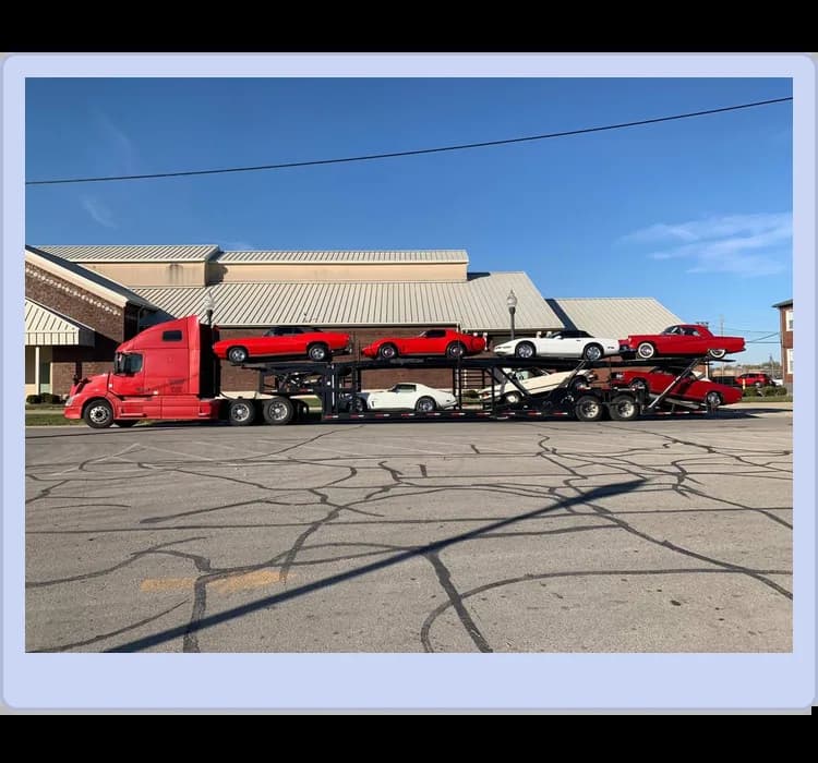 A red car carrier truck is parked, loaded with several red sports cars on both its upper and lower levels. Buildings and a clear sky are in the background.